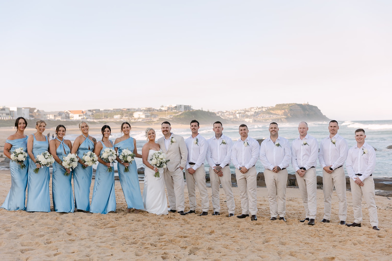 The wedding party on the sand with the headland behind. Jasmin & Jye's wedding at Merewether Surfhouse & Ocean Baths, Newcastle